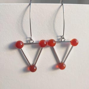 May include: A pair of geometric earrings featuring a triangular design. Each earring has a silver-colored metal frame with three red, spherical beads at each point. The earrings hang from thin, curved silver wires.