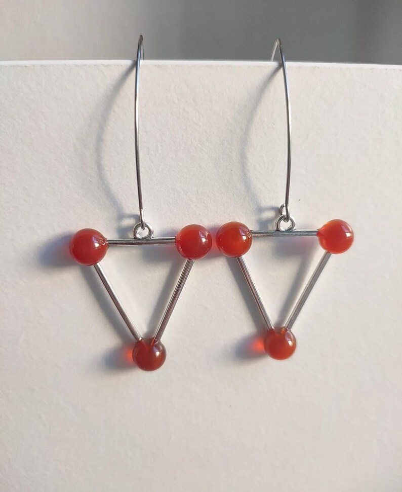 May include: A pair of geometric earrings featuring a triangular design. Each earring has a silver-colored metal frame with three red, spherical beads at each point. The earrings hang from thin, curved silver wires.