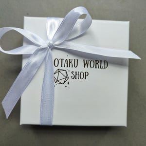 May include: A white gift box tied with a light blue satin ribbon. The box has the text "OTAKU WORLD SHOP" printed in black, along with a geometric design. The box is on a gray surface.