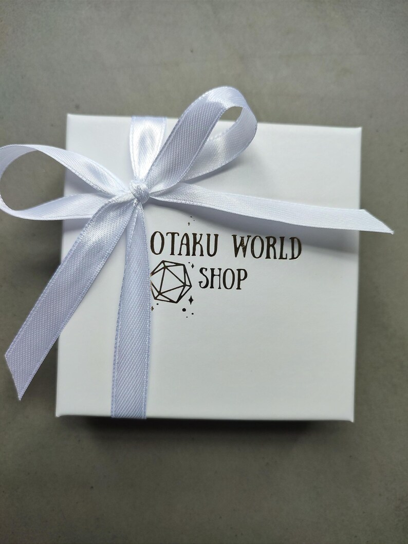 May include: A white gift box tied with a light blue satin ribbon. The box has the text "OTAKU WORLD SHOP" printed in black, along with a geometric design. The box is on a gray surface.