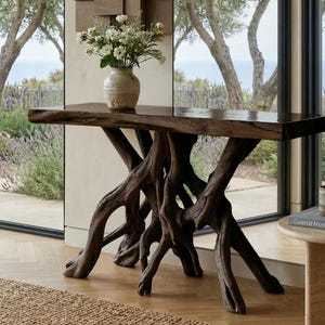 May include: A dark brown, live-edge console table with a natural wood root base. The table is topped with a vase of white flowers. A small side table with a mug and a book is to the right.