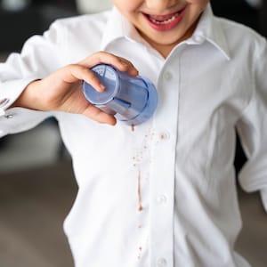 Puede incluir: Una persona joven que lleva una camisa blanca con una mancha marrón en la parte delantera. La persona sostiene una taza de plástico azul en su mano.