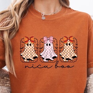 May include: A burnt orange t-shirt featuring three cartoon ghosts with bows and checkered patterns. The text "nicu boo" is written in a cursive font below the ghosts. The shirt is a YAM COLOR.