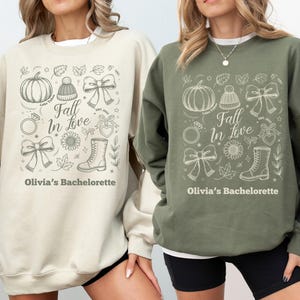 May include: Two sweatshirts, one beige and one olive green, each with a fall-themed graphic. The graphic includes a pumpkin, bow, ring, boot, and the text "Fall In Love" and "Olivia's Bachelorette".