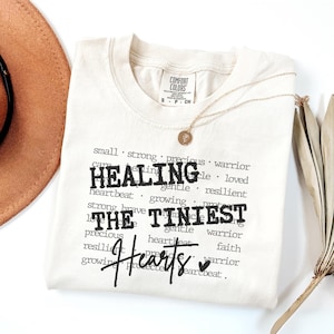 May include: Cream-colored t-shirt with the words "HEALING THE TINIEST HEARTS" in black. A gold necklace with a circular pendant rests on the shirt. A brown hat and dried palm leaf are also in the image.