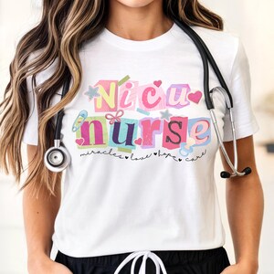 May include: A white t-shirt featuring a colorful graphic with the text "NICU nurse" in block letters, surrounded by small illustrations of a safety pin, hearts, stars, and a pacifier, with the words "miracles love hope care" written below.