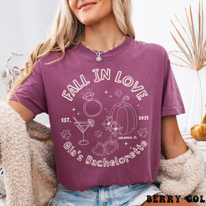 以下が含まれることがあります： ベリー色のTシャツ。白で「FALL IN LOVE」と「Gia's Bachelorette」の文字。デザインには指輪、カボチャ、カクテル、ハートが含まれています。快適なフィット感のシャツです。