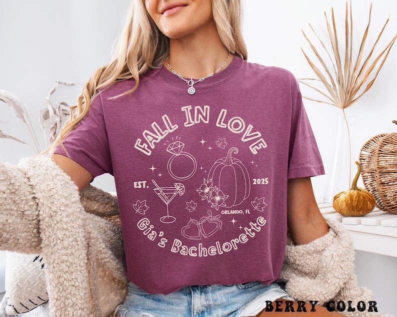 以下が含まれることがあります： ベリー色のTシャツ。白で「FALL IN LOVE」と「Gia's Bachelorette」の文字。デザインには指輪、カボチャ、カクテル、ハートが含まれています。快適なフィット感のシャツです。