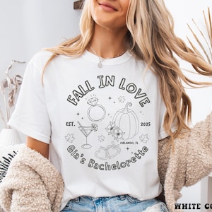 以下が含まれることがあります： 黒いグラフィックデザインの白いTシャツ。デザインには、ダイヤモンドの指輪、カクテル、カボチャ、花の要素の上に「FALL IN LOVE」というテキストが含まれています。「Gia's Bachelorette」と「2025」の文字も含まれています。