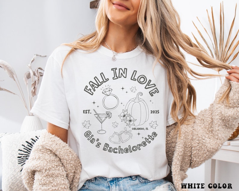以下が含まれることがあります： 黒いグラフィックデザインの白いTシャツ。デザインには、ダイヤモンドの指輪、カクテル、カボチャ、花の要素の上に「FALL IN LOVE」というテキストが含まれています。「Gia's Bachelorette」と「2025」の文字も含まれています。