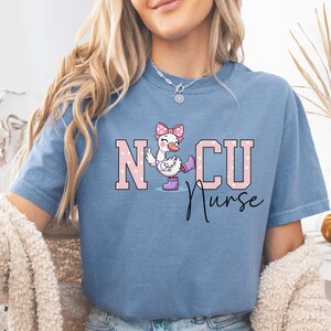 May include: A blue t-shirt featuring a graphic of a white duck wearing a pink bow, purple boots, and a stethoscope. The text NICU Nurse is printed in large, light pink, white-dotted block letters with the word Nurse in black script below.