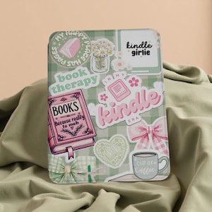 May include: A Kindle case with a green and white check pattern, decorated with stickers. The stickers feature a pink book, daisies, a bow, and the phrases "book therapy", "Kindle girlie", and "Cup of Coffee".