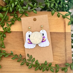 Peut inclure: Une paire de boucles d'oreilles pendantes en forme de fant&ocirc;me blanc avec des attaches dor&eacute;es. Chaque fant&ocirc;me tient un petit livre violet avec un croissant de lune jaune. Une pi&egrave;ce de 25 cents est pos&eacute;e sur les bijoux pour montrer l'&eacute;chelle.