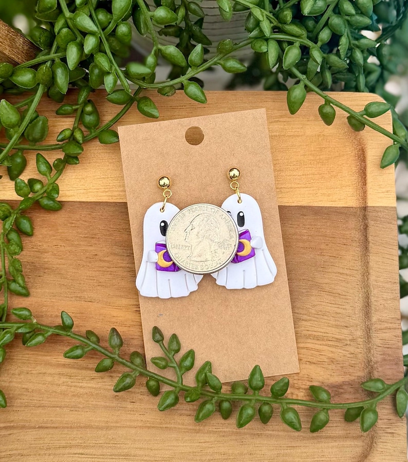 Peut inclure: Une paire de boucles d'oreilles pendantes en forme de fant&ocirc;me blanc avec des attaches dor&eacute;es. Chaque fant&ocirc;me tient un petit livre violet avec un croissant de lune jaune. Une pi&egrave;ce de 25 cents est pos&eacute;e sur les bijoux pour montrer l'&eacute;chelle.