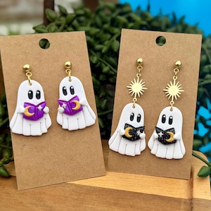 May include: Two pairs of dangle earrings on brown cardboard display cards. One pair features white ghosts holding purple books with gold crescent moons. The other pair features white ghosts holding black books with gold sun and moon charms.