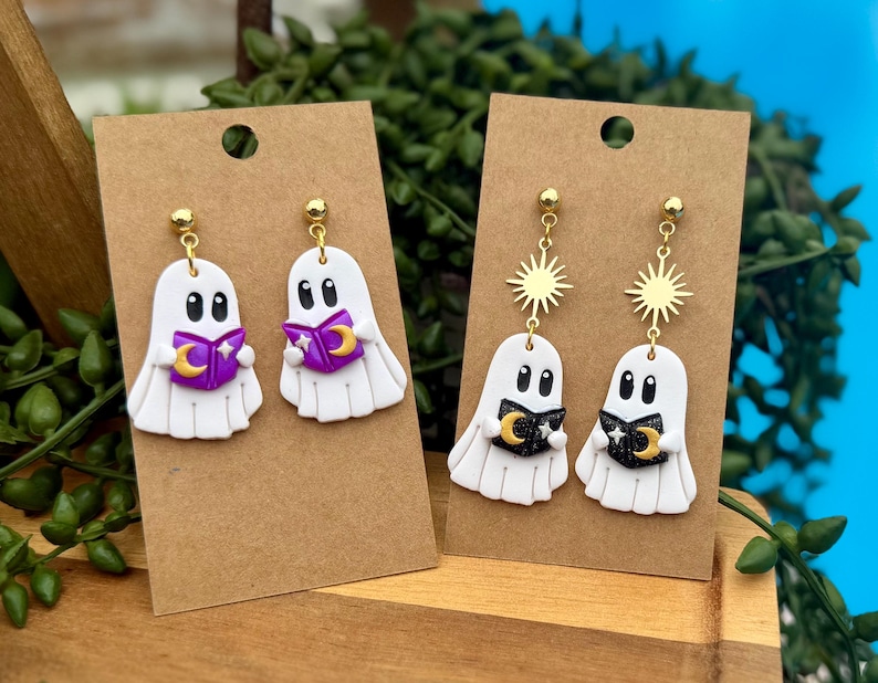 Peut inclure: Deux paires de boucles d'oreilles pendantes sur des supports en carton brun. Une paire repr&eacute;sente des fant&ocirc;mes blancs tenant des livres violets avec des croissants de lune dor&eacute;s. L'autre paire montre des fant&ocirc;mes blancs avec des livres noirs.