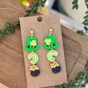 May include: A pair of green, gold, and black dangle earrings featuring a leprechaun hat, rainbow, and pot of gold charms.