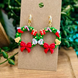 May include: A pair of festive Christmas wreath earrings. Each green wreath features red bows, candy canes, snowmen, and gift boxes. Gold-colored hoops attach the earrings to a brown card.