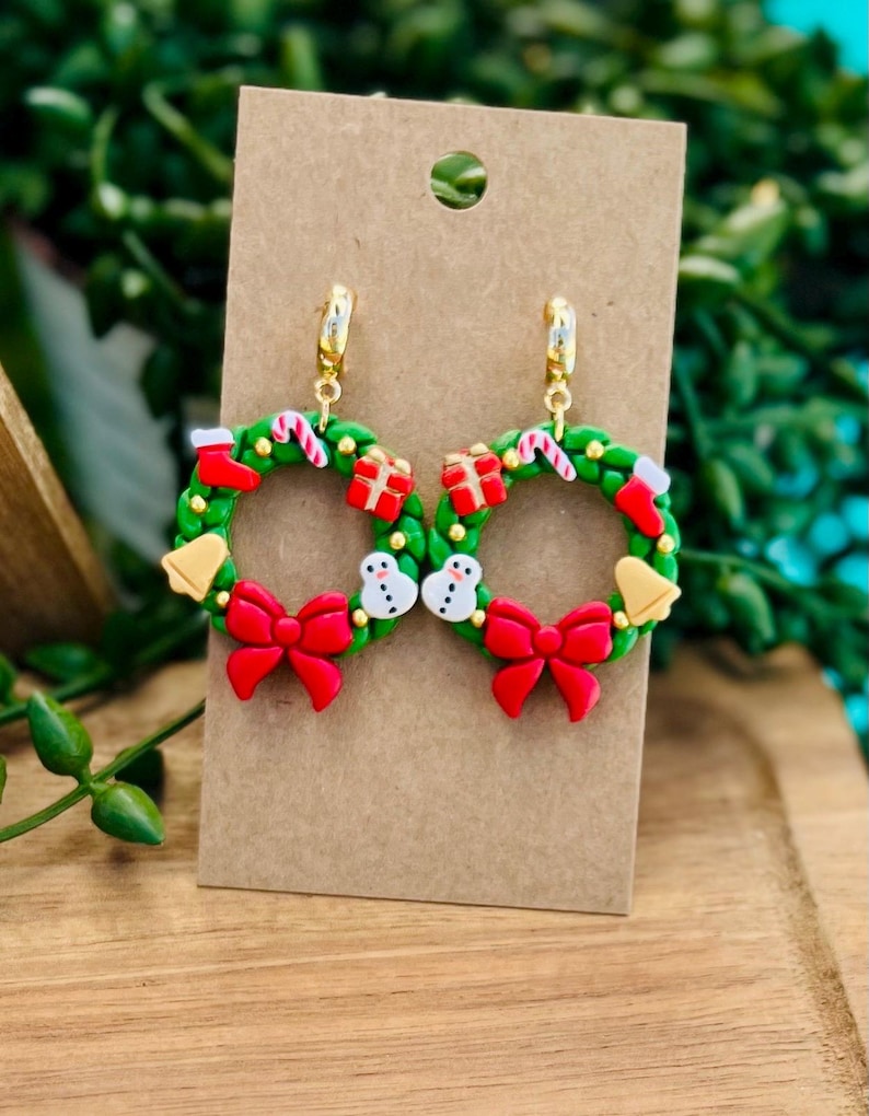 May include: A pair of festive Christmas wreath earrings. Each green wreath features red bows, candy canes, snowmen, and gift boxes. Gold-colored hoops attach the earrings to a brown card.