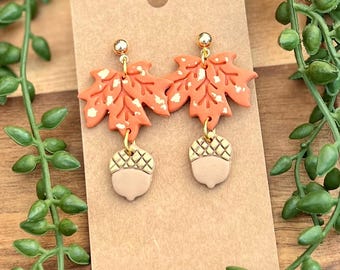 May include: A pair of orange and gold dangle earrings featuring a maple leaf design with white speckles and a small acorn charm.