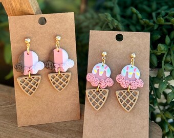 Puede incluir: Dos pares de pendientes colgantes con forma de conos de helado. Un par presenta bolas marrones, rosas y blancas con conos de barquillo. El otro par presenta bolas rosas con glaseado blanco, virutas de colores y conos de barquillo. Todos tienen postes dorados.
