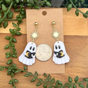 Peut inclure: Une paire de boucles d'oreilles fant&ocirc;mes blanches avec des breloques soleil dor&eacute;es et des livres noirs orn&eacute;s d'un croissant de lune et d'une &eacute;toile jaunes. Les boucles d'oreilles sont pr&eacute;sent&eacute;es sur une carte brune &agrave; c&ocirc;t&eacute; d'une pi&egrave;ce de monnaie.