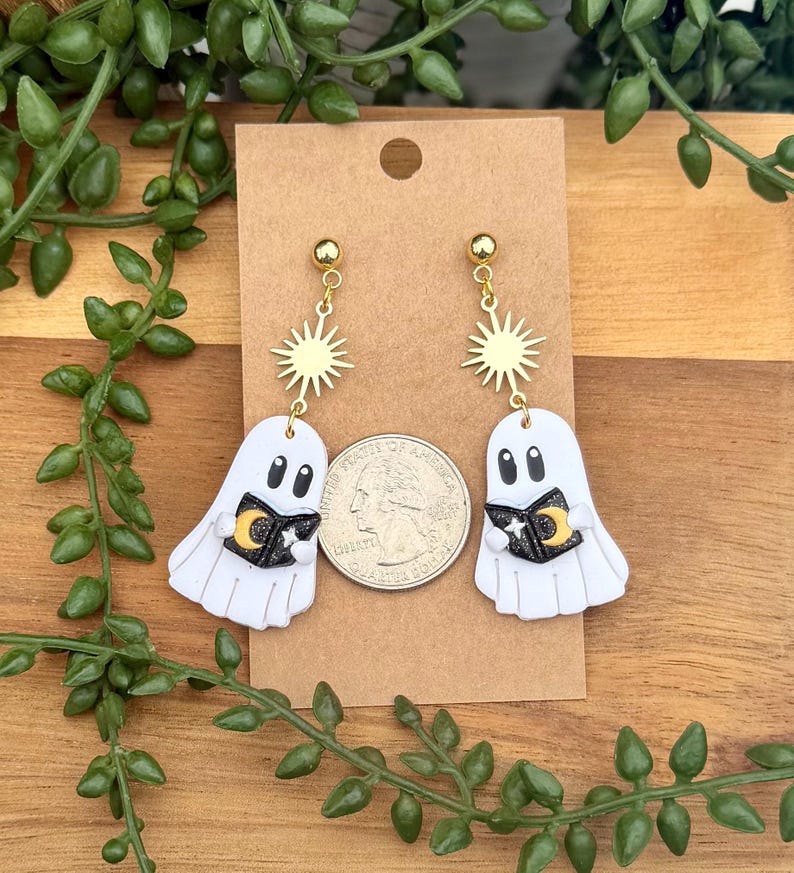 Peut inclure: Une paire de boucles d'oreilles fant&ocirc;mes blanches avec des breloques soleil dor&eacute;es et des livres noirs orn&eacute;s d'un croissant de lune et d'une &eacute;toile jaunes. Les boucles d'oreilles sont pr&eacute;sent&eacute;es sur une carte brune &agrave; c&ocirc;t&eacute; d'une pi&egrave;ce de monnaie.