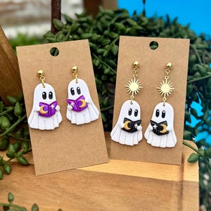 Peut inclure: Deux paires de boucles d'oreilles fant&ocirc;mes blanches sur des supports en carton brun. La premi&egrave;re paire montre des fant&ocirc;mes tenant des livres violets avec des lunes dor&eacute;es. La seconde paire pr&eacute;sente des soleils dor&eacute;s et des livres noirs avec des lunes.
