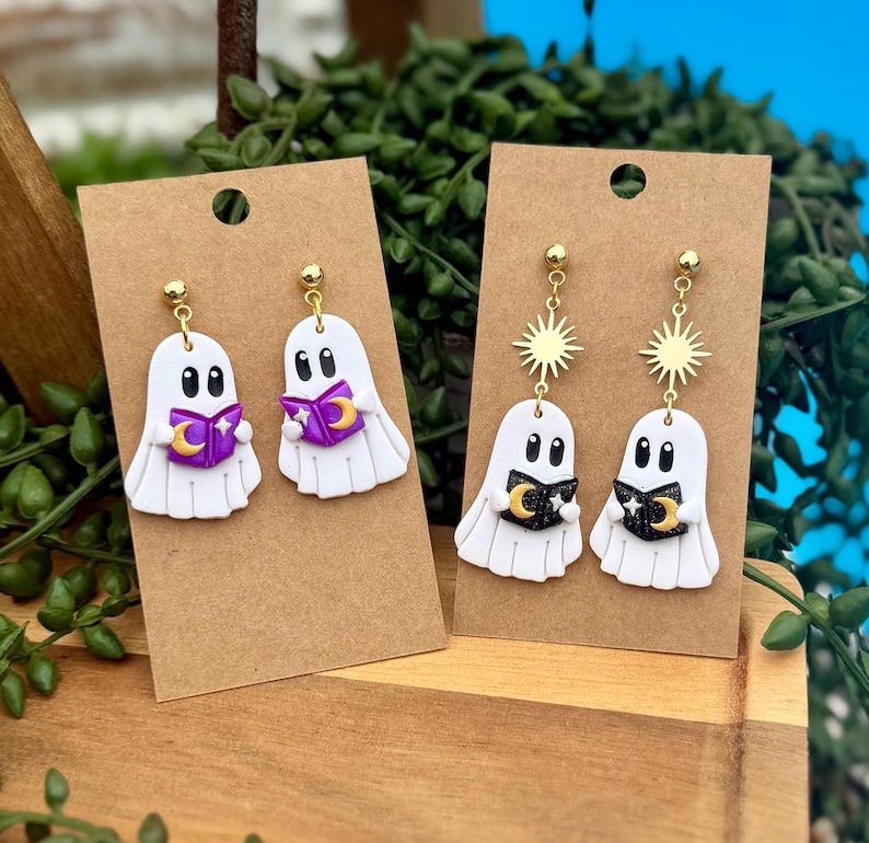 Peut inclure: Deux paires de boucles d'oreilles fant&ocirc;mes blanches sur des supports en carton brun. La premi&egrave;re paire montre des fant&ocirc;mes tenant des livres violets avec des lunes dor&eacute;es. La seconde paire pr&eacute;sente des soleils dor&eacute;s et des livres noirs avec des lunes.