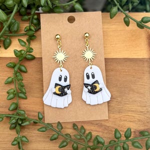 Peut inclure: Une paire de boucles d'oreilles pendantes repr&eacute;sentant des fant&ocirc;mes blancs tenant de petits livres noirs avec des d&eacute;tails de croissant de lune dor&eacute; et d'&eacute;toile blanche. Chaque fant&ocirc;me est suspendu &agrave; un motif soleil dor&eacute; et un clou d'oreille.