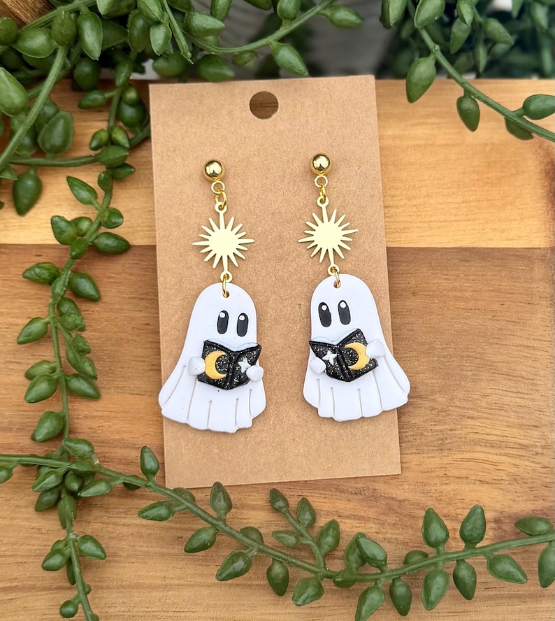 Peut inclure: Une paire de boucles d'oreilles pendantes repr&eacute;sentant des fant&ocirc;mes blancs tenant de petits livres noirs avec des d&eacute;tails de croissant de lune dor&eacute; et d'&eacute;toile blanche. Chaque fant&ocirc;me est suspendu &agrave; un motif soleil dor&eacute; et un clou d'oreille.