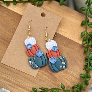 May include: A pair of handmade earrings featuring a stacked pumpkin design. Each earring has three pumpkins in white, orange, and teal, adorned with blue flower accents and gold leaf details. The earrings hang from gold-colored hooks.
