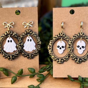 Op de afbeelding: Twee paar Halloween-oorbellen. Eén paar heeft witte spookbedels, de andere witte schedelbedels. Beide sets zijn omlijst in sierlijk goudkleurig filigraan. De oorbellen hangen aan goudkleurige haken en worden gepresenteerd op bruin karton.