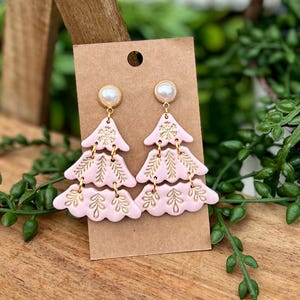 Puede incluir: Pendientes rosas de árbol de Navidad con detalles de hojas doradas y tachuelas con forma de perla. Los pendientes están hechos de un material suave, similar a la arcilla, y están sujetos a una tarjeta marrón.