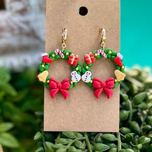 May include: A pair of festive Christmas wreath earrings. Each earring features a green wreath adorned with red bows, gift boxes, candy canes, snowmen, and bells. The earrings are attached to gold-colored hoops and are displayed on a brown card.