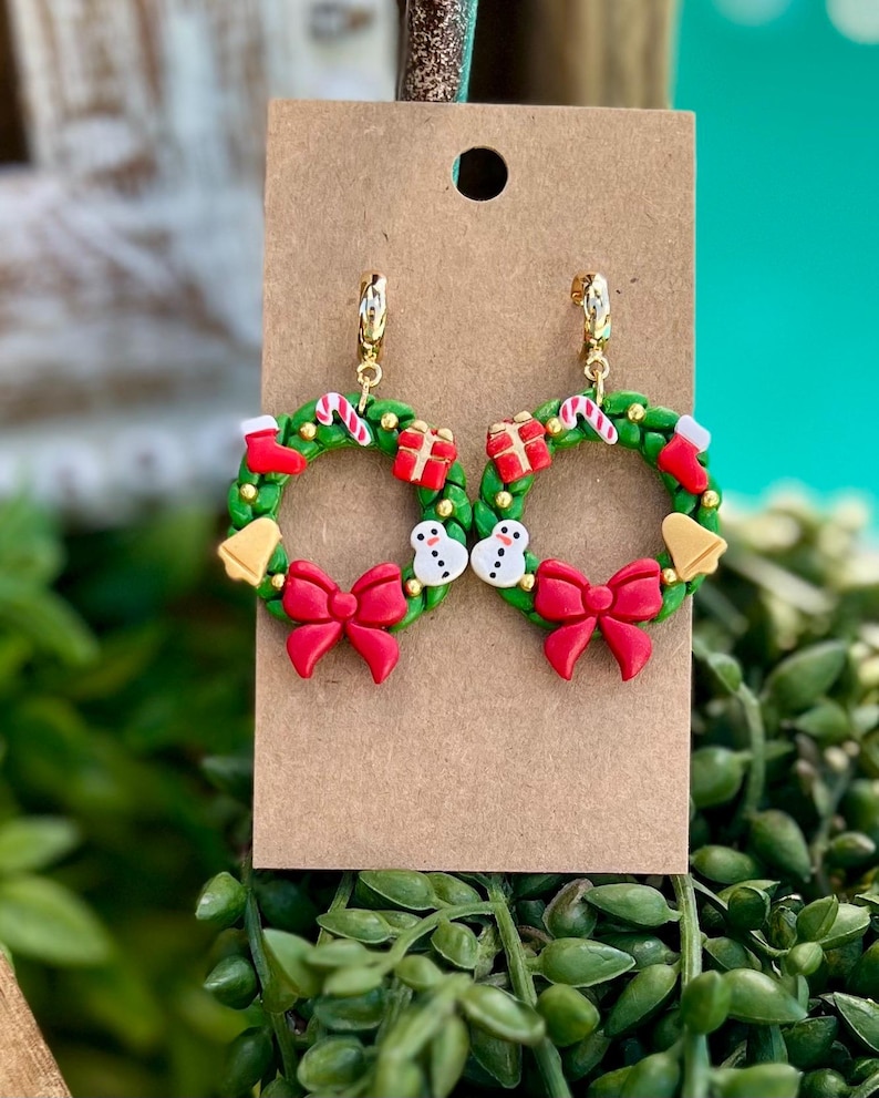 May include: A pair of festive Christmas wreath earrings. Each earring features a green wreath adorned with red bows, gift boxes, candy canes, snowmen, and bells. The earrings are attached to gold-colored hoops and are displayed on a brown card.