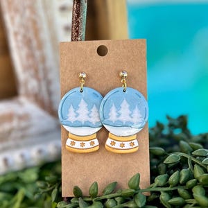 May include: A pair of blue and white snow globe earrings. The earrings feature a light blue background with white trees and a white base with gold star details. The earrings are attached to a brown card with a gold-colored metal hook.