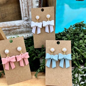 Puede incluir: Tres pares de pendientes en forma de lazo con detalles de perlas. Los pendientes se muestran en cartulina marrón. Los lazos son blancos, rosas y azul claro.