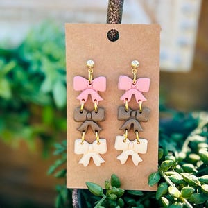 Puede incluir: Un par de pendientes colgantes con tres lazos superpuestos. El lazo superior es rosa, el lazo del medio es marrón y el lazo inferior es blanco.