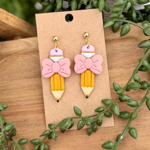 May include: A pair of novelty earrings shaped like yellow pencils with pink bows. The pencil earrings have gold-colored studs and are displayed on a brown card. The earrings are a fun accessory for teachers or students.