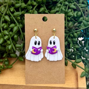 Peut inclure: Une paire de boucles d'oreilles pendantes en forme de fant&ocirc;me blanc avec des yeux noirs, tenant un livre violet orn&eacute; d'un croissant de lune et d'une &eacute;toile dor&eacute;s. Les boucles sont fix&eacute;es sur un support en carton brun avec des clous dor&eacute;s.