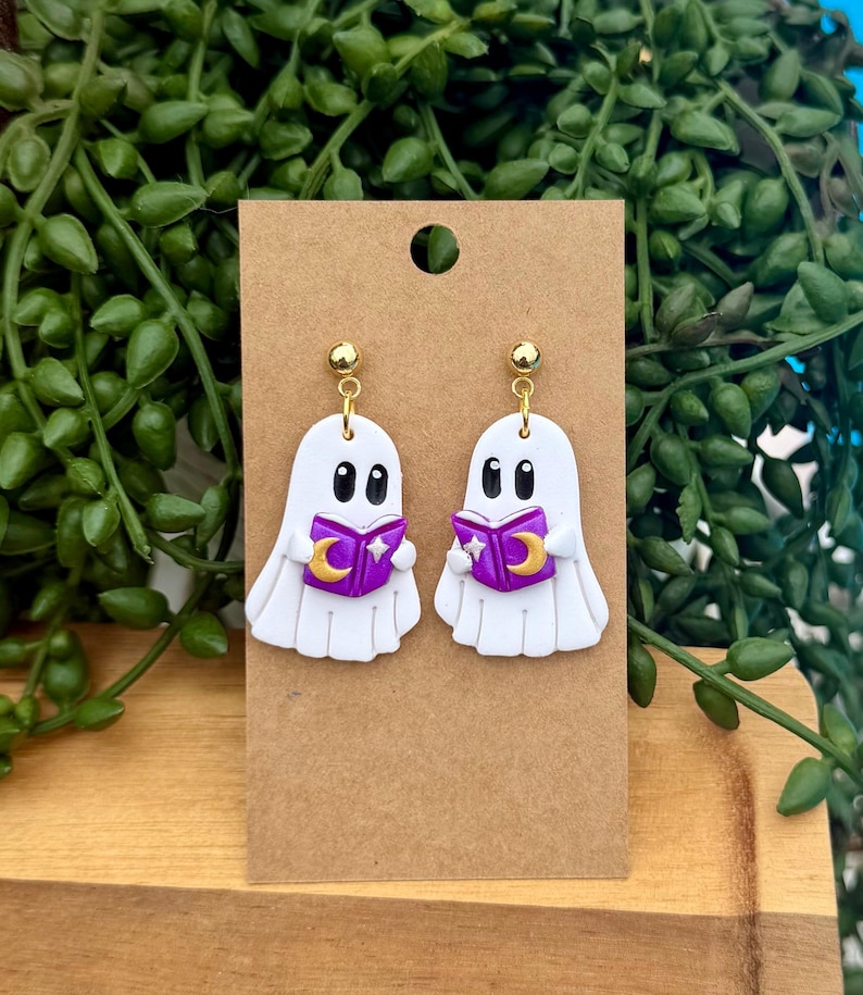 Peut inclure: Une paire de boucles d'oreilles pendantes en forme de fant&ocirc;me blanc avec des yeux noirs, tenant un livre violet orn&eacute; d'un croissant de lune et d'une &eacute;toile dor&eacute;s. Les boucles sont fix&eacute;es sur un support en carton brun avec des clous dor&eacute;s.