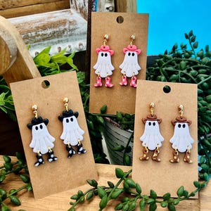 Op de afbeelding: Handgemaakte Halloween-oorbellen met witte spookfiguren met hoeden en laarzen. De oorbellen worden gepresenteerd op bruine oorbellenkaarten. De hoeden en laarzen zijn zwart, roze en bruin. De oorbellen hebben goudkleurige pinnen.