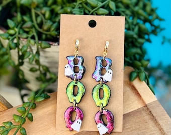 May include: A pair of Halloween-themed dangle earrings spelling out "BOO". The letters are iridescent with a glittery finish, featuring small white ghost charms. Gold-colored ear wires complete the design. The earrings are displayed on a brown card.