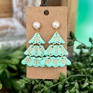 May include: Mint green Christmas tree earrings with gold accents. Each earring features three tiers of tree shapes with gold leaf and snowflake details. The earrings are attached to a gold-colored stud with a pearl-like bead.