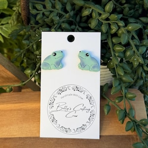 May include: Two light green frog earrings with a light blue outline and black eyes, displayed on a white card. The card features the text "Handmade With Love" and "Betty's Crafting Crew" in a circular design.