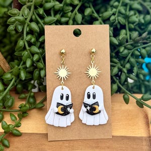 Peut inclure: Une paire de boucles d'oreilles pendantes avec des breloques en forme de fant&ocirc;me blanc tenant de petits livres noirs orn&eacute;s d'un croissant de lune et d'&eacute;toiles dor&eacute;s. Chaque fant&ocirc;me est suspendu &agrave; un connecteur soleil dor&eacute; sur un clou d'oreille.
