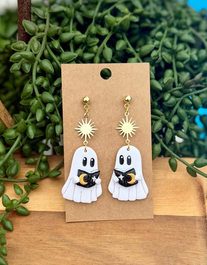 Peut inclure: Une paire de boucles d'oreilles pendantes avec des breloques en forme de fant&ocirc;me blanc tenant de petits livres noirs orn&eacute;s d'un croissant de lune et d'&eacute;toiles dor&eacute;s. Chaque fant&ocirc;me est suspendu &agrave; un connecteur soleil dor&eacute; sur un clou d'oreille.