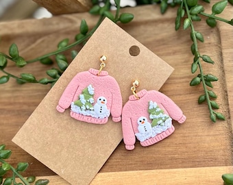 May include: A pair of pink polymer clay earrings shaped like Christmas sweaters with a snowman and a pine tree design.