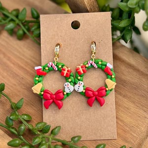May include: A pair of festive Christmas wreath earrings. Each earring features a green wreath adorned with red bows, candy canes, gift boxes, snowmen, and gold bells. The earrings are attached to gold-colored ear wires and displayed on a brown card.
